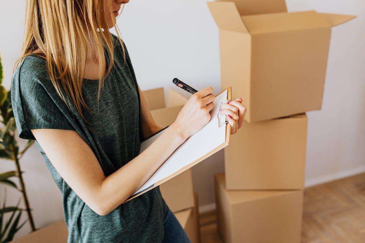 A young woman writes on a clipboard while organizing moving boxes in her home.