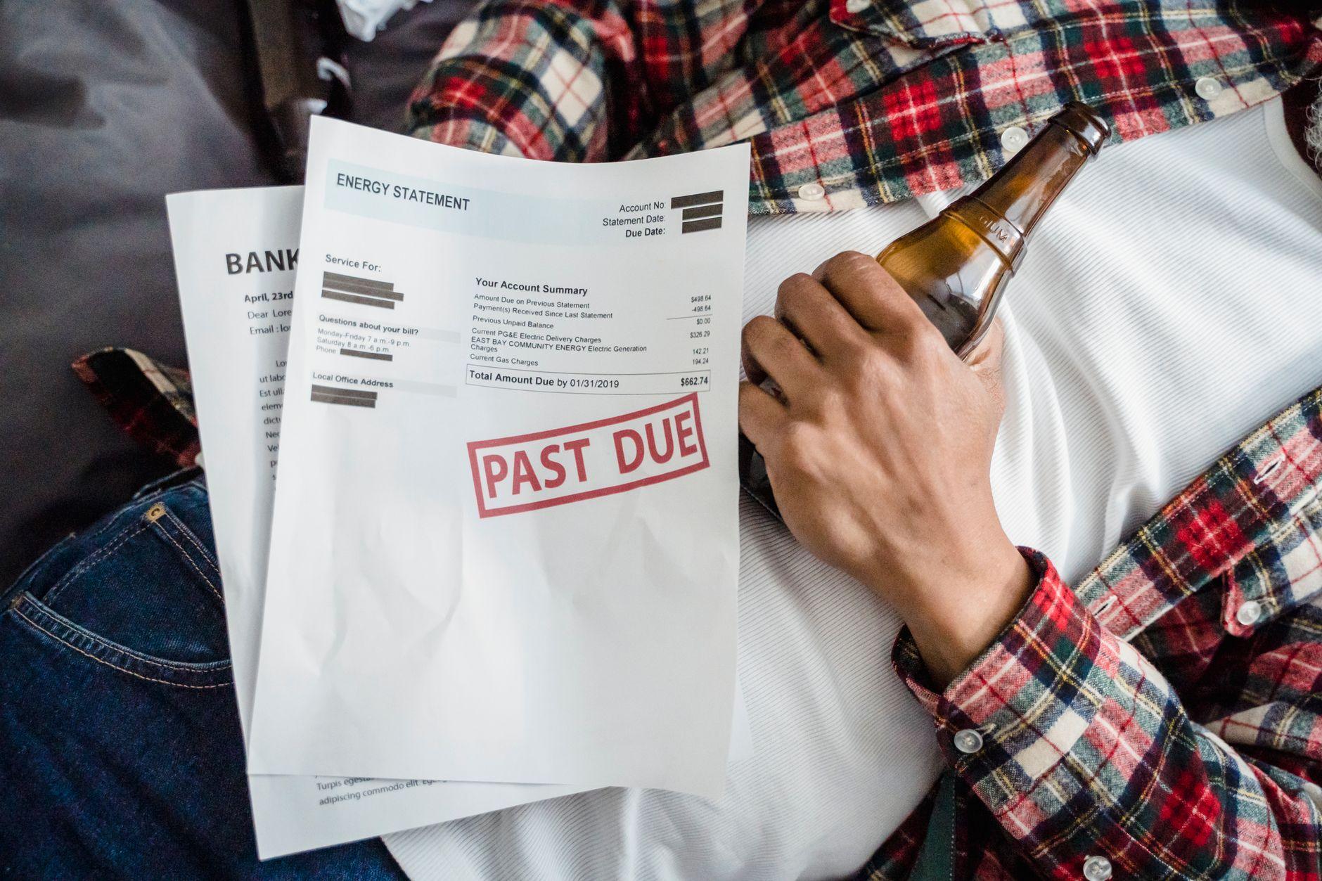A man relaxes with a beer bottle while holding past due bills, highlighting financial stress.